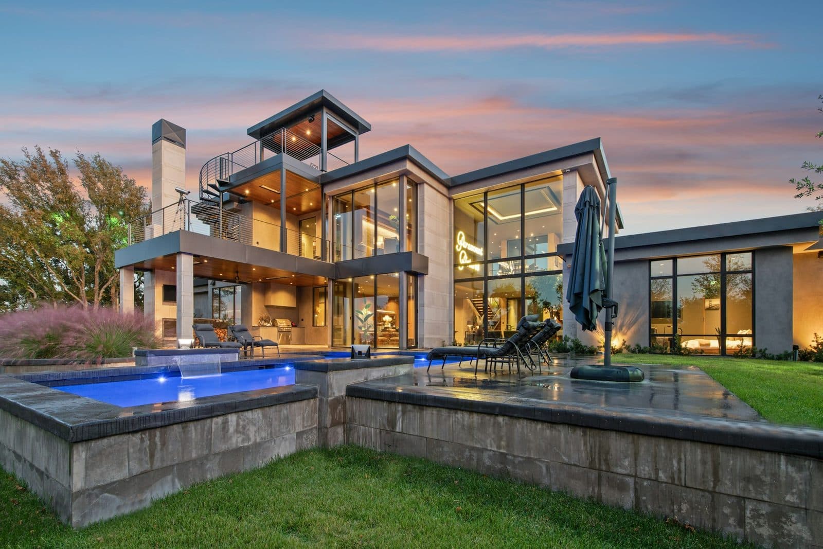 Dusk backyard with a glowing pool, stone deck, green lawn, and covered lounge